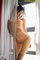 A woman in a nude bodysuit leaning against a window.